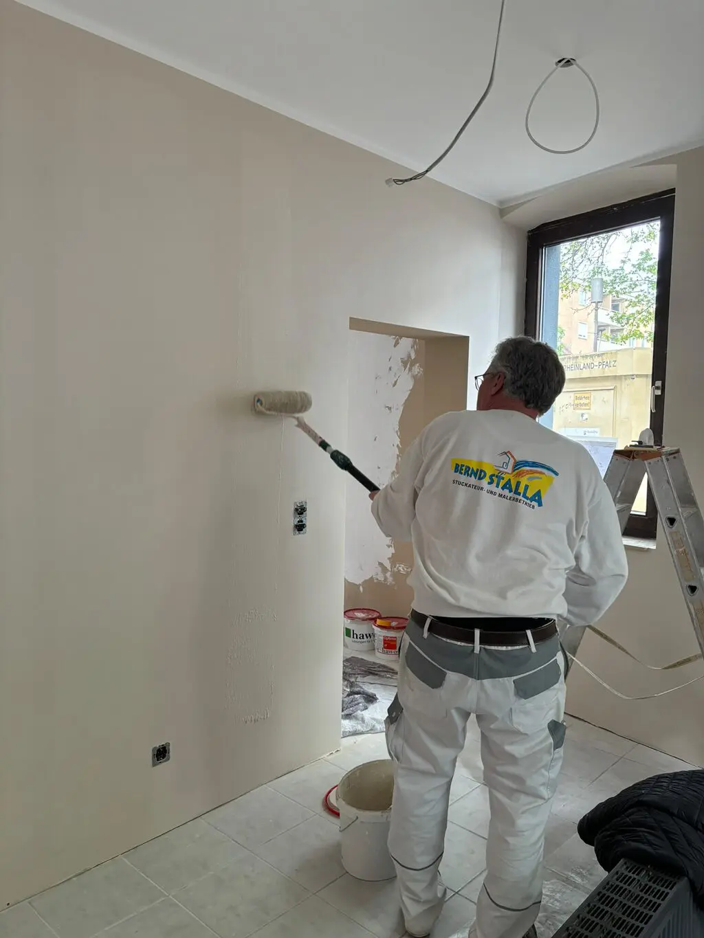 Maler mit weißer Arbeitskleidung streicht Wand mit Farbrolle in hellem Raum mit Fenster und Leiter