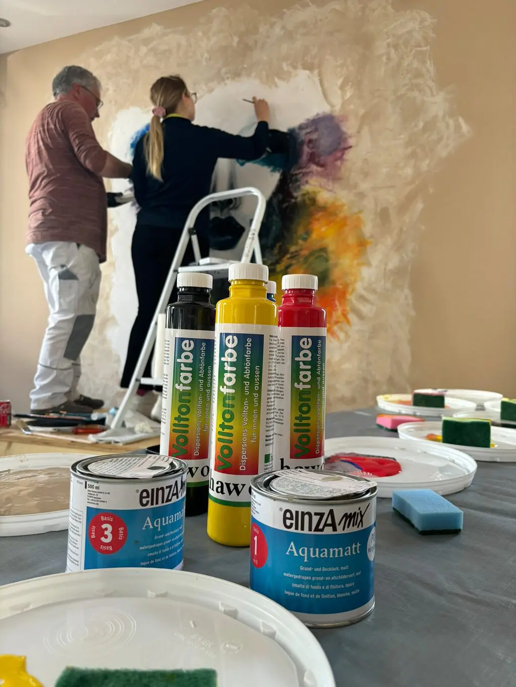 Zwei Personen, eine auf einer Leiter stehend, malen gemeinsam ein Wandgemälde mit bunten Farben auf einer hellen Wand
