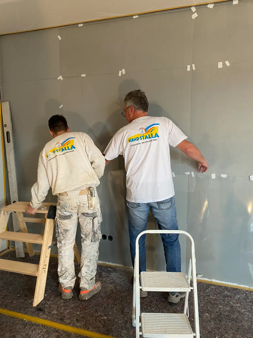 Zwei Männer mit weißen T-Shirts und Arbeitskleidung messen eine Wand ab, eine kleine Leiter steht davor
