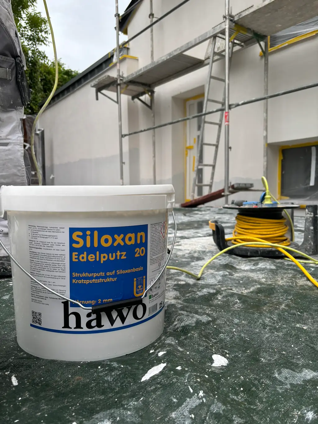 Nahaufnahme eines Farbeimers mit der Aufschrift 'Siloxan Edelputz 20' vor einer Baustelle mit Gerüst und Kabelrolle