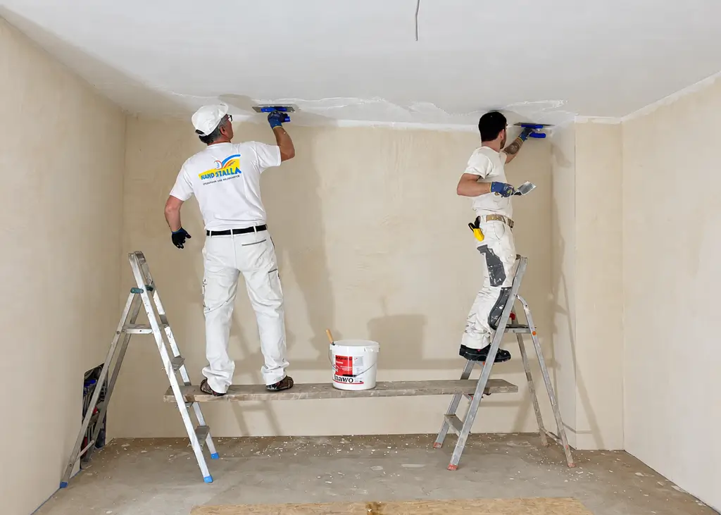 Zwei Handwerker in weißer Arbeitskleidung tragen Deckenputz in einem Raum auf, sie stehen auf einer Arbeitsbühne zwischen zwei Leitern