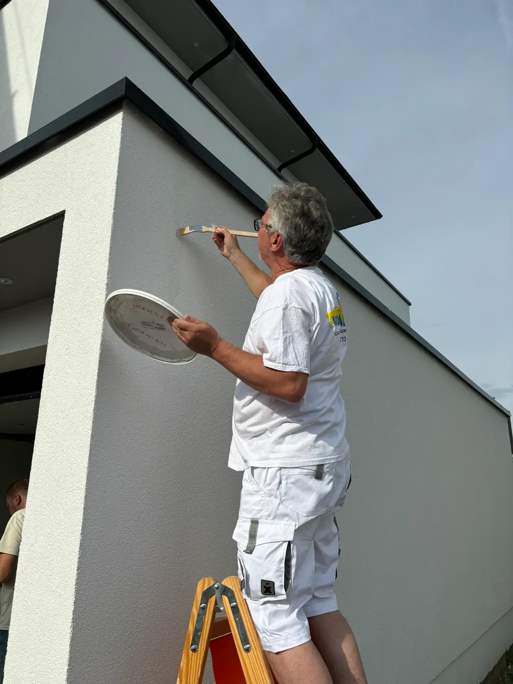 Mann auf Leiter streicht Fassade mit Pinsel und Farbpalette