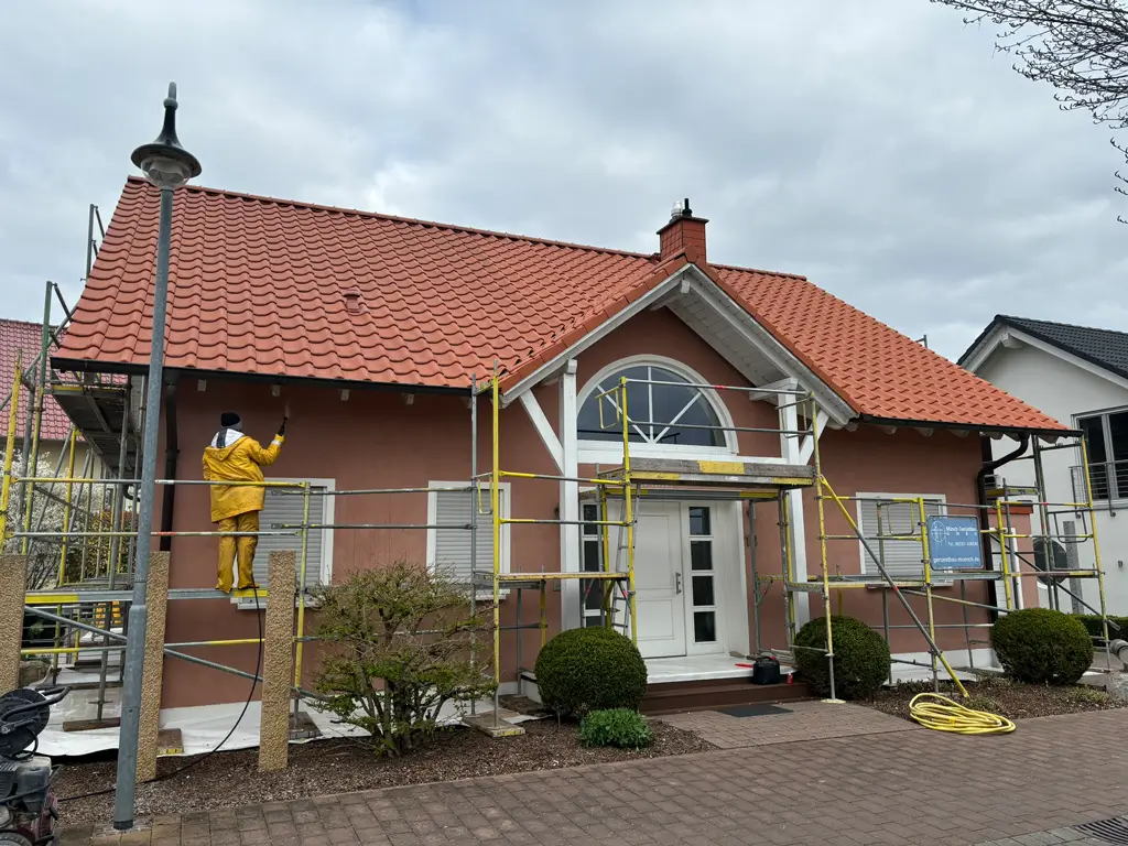 Haus mit rotem Ziegeldach und Gerüst davor, auf dem eine Person in gelber Arbeitskleidung die Fassade streicht