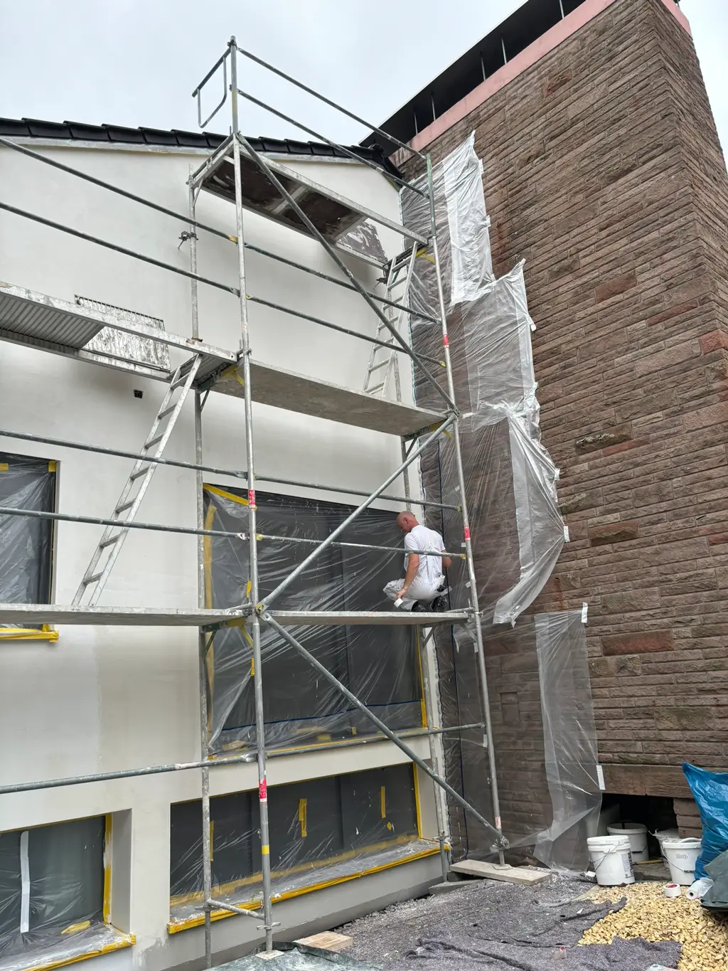 Gerüst vor einer weißen Hausfassade mit einem Mann, der an der Wand arbeitet, neben einer Fassade mit Steinverkleidung