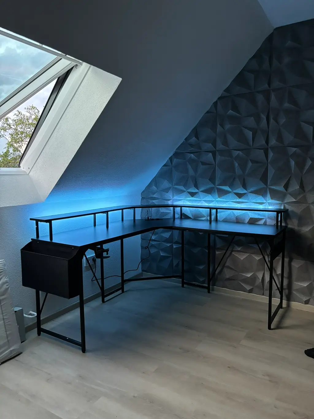 Eckschreibtisch mit schwarzem Metallgestell vor einer Wand mit 3D-Struktur und blauem LED-Licht unter einem Dachfenster