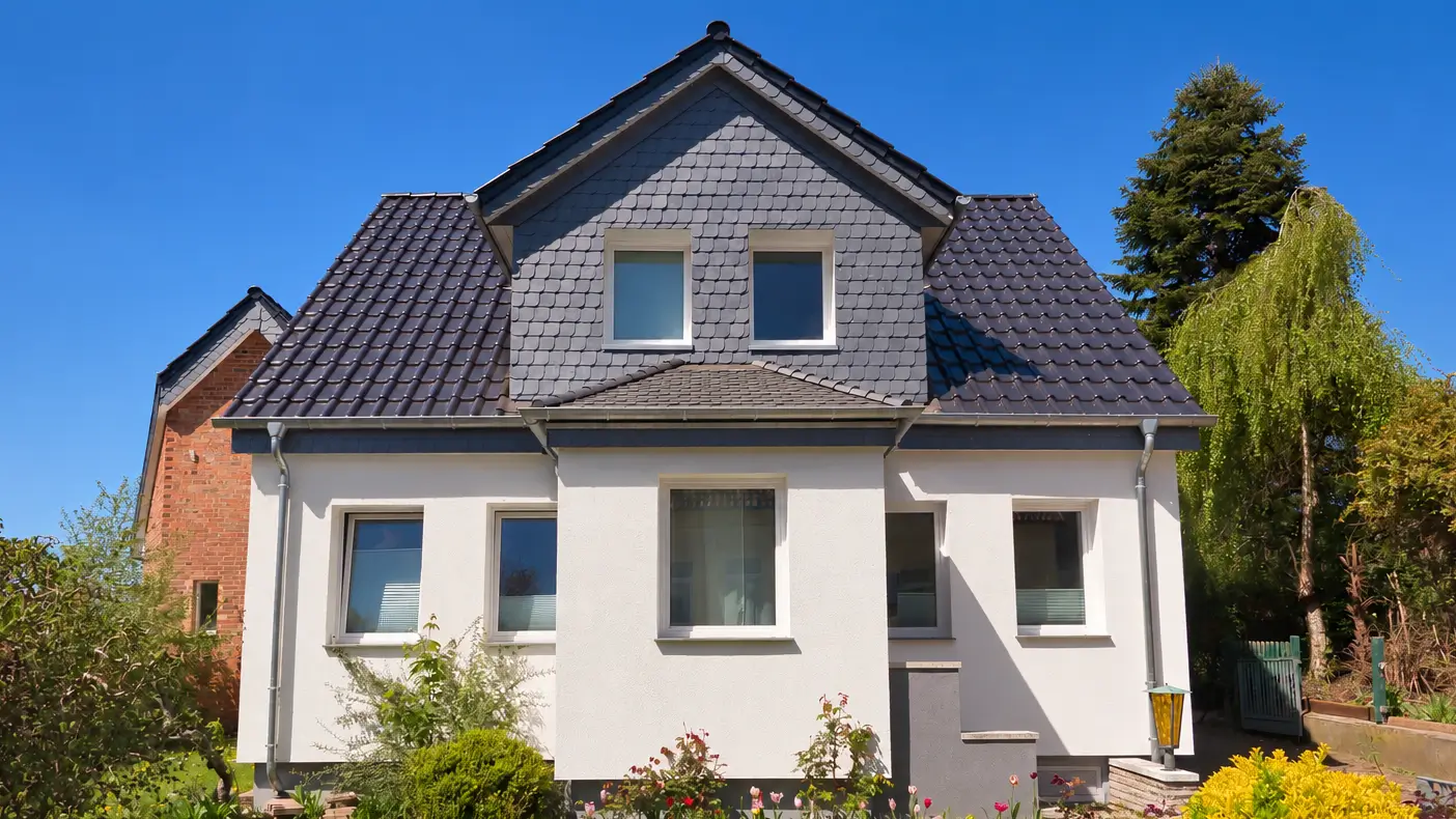 Haus nach Renovierung Zweistöckiges Einfamilienhaus mit weißer Fassade, grauem Satteldach und mehreren Fenstern, umgeben von Büschen und Bäumen