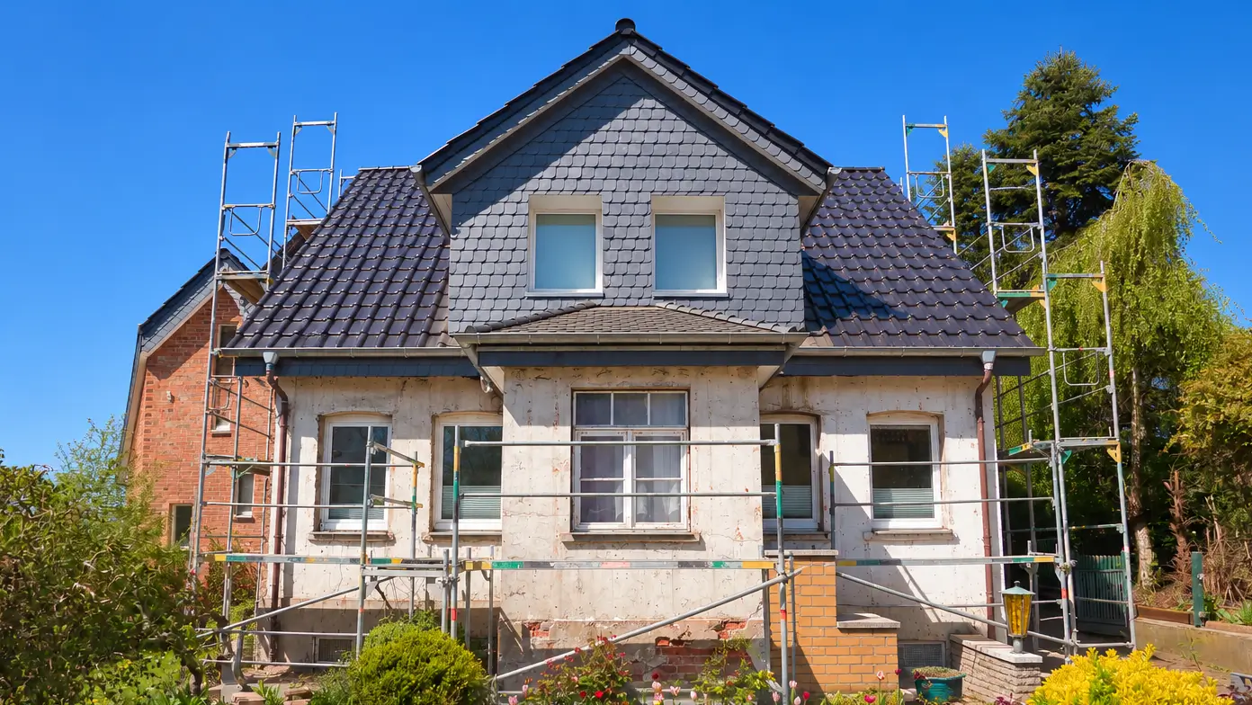 Haus während Renovierung Ein zweistöckiges Haus mit dunklem Dach und eingerüsteter Fassade vor blauem Himmel