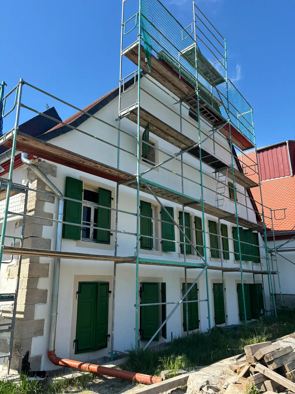 Hausfassade mit grünen Fensterläden und Gerüst vor weißer Wand unter blauem Himmel