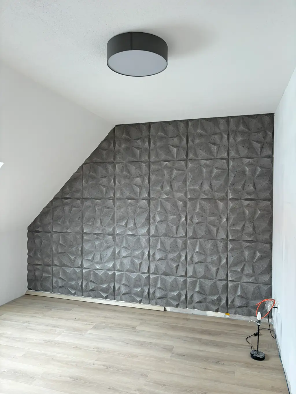 Wand mit grauen, dreidimensional strukturierten Schallschutzplatten in einem leeren Raum mit hellem Holzboden und runder Deckenleuchte