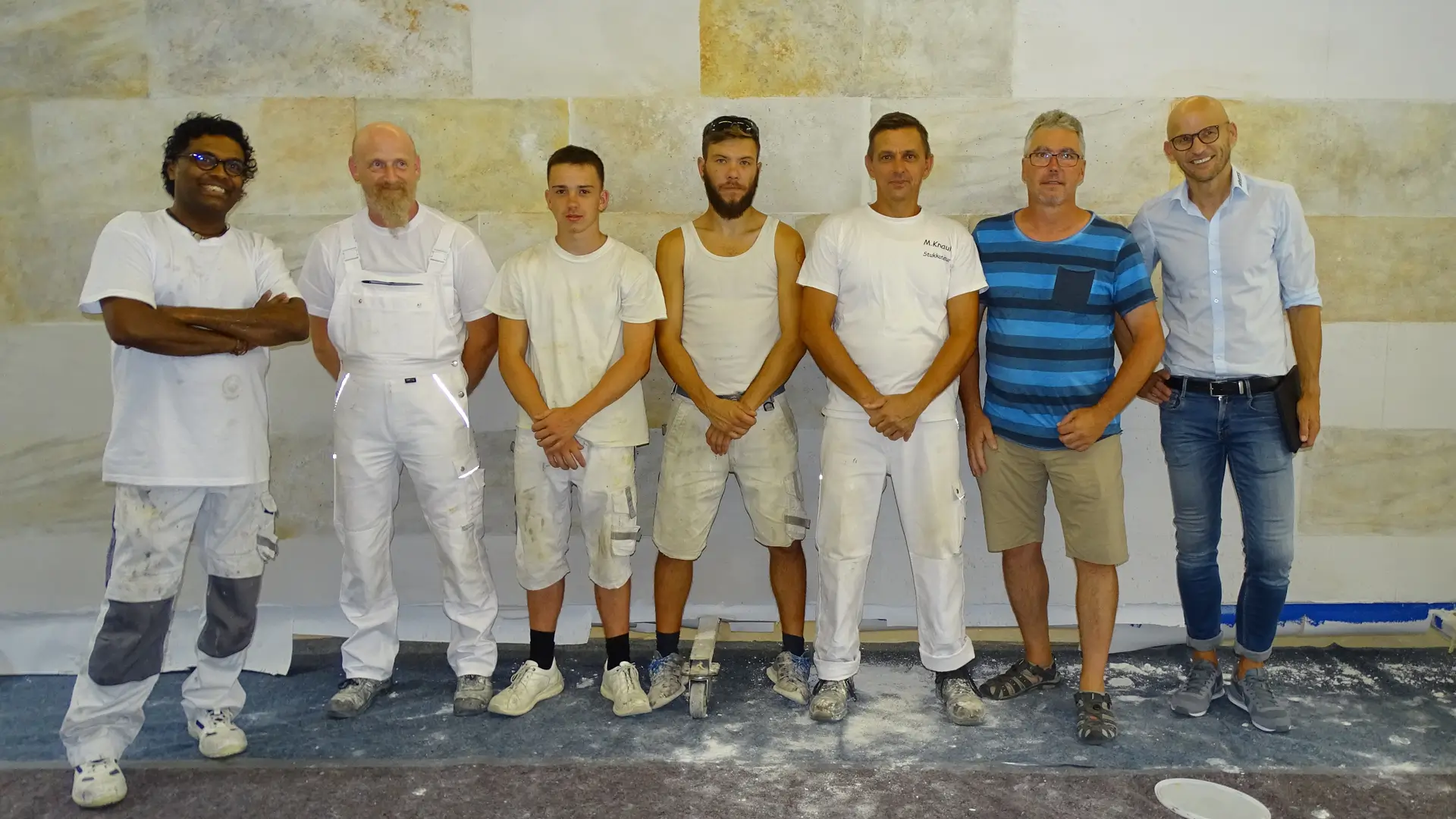 Team Sieben Männer in Arbeitskleidung und Freizeitkleidung stehen nebeneinander vor einer hellen Wand in einem Raum mit Malerbedarf auf dem Boden