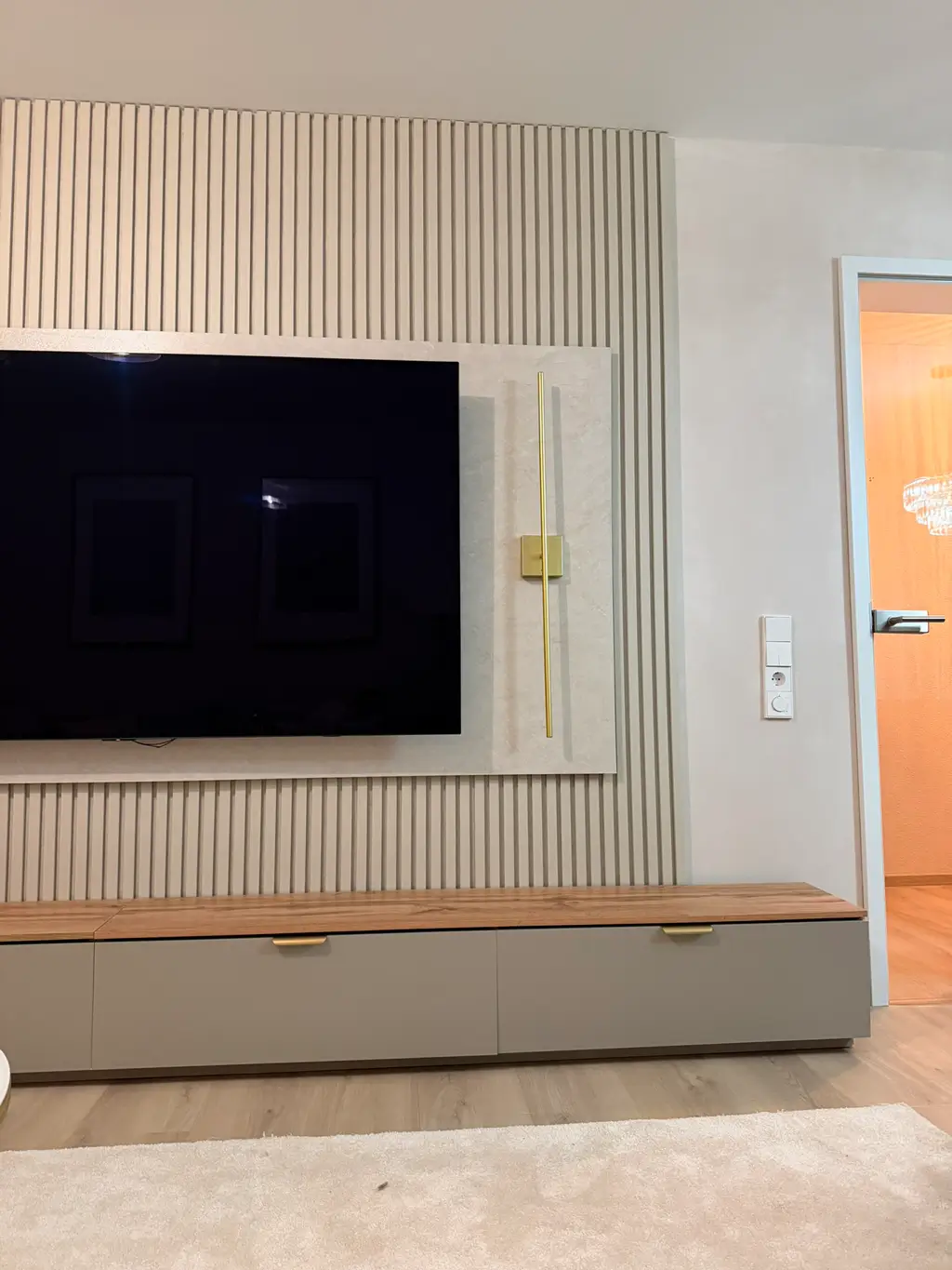 Flacher Fernseher an einer vertikal gerillten Trockenbauwand über einem niedrigen Sideboard mit Holzdeckel neben einer offenen Tür zu einem hellen Raum
