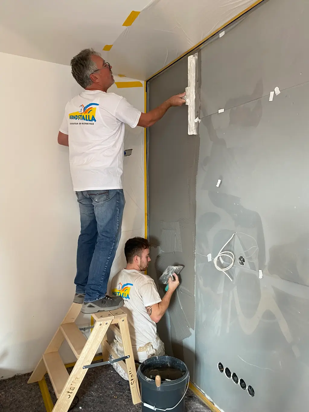 Zwei Männer in weißen T-Shirts mit Logo verputzen eine Wand, einer steht auf einer Leiter und trägt Putz mit einer Glättkelle auf, der andere hält eine Kelle und steht am Boden neben einem Eimer.