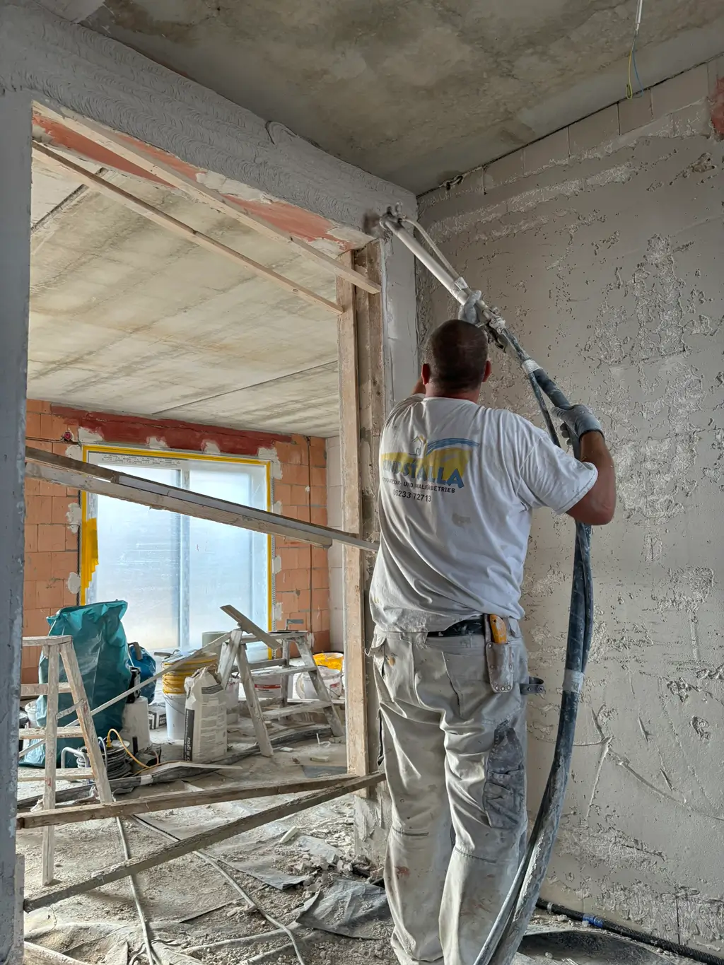 Handwerker mit weißer Arbeitskleidung verputzt mit einer Spritzpistole die Wand um einen Türrahmen in einem Rohbau