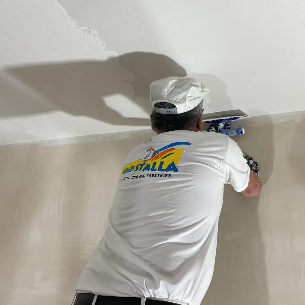 Handwerker mit weißem T-Shirt und Mütze verputzt Decke mit Glättkelle
