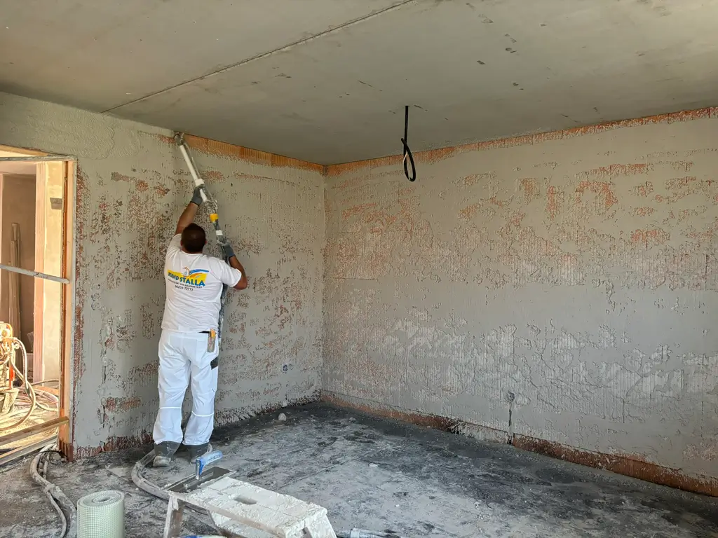 Handwerker in weißer Arbeitskleidung trägt Putz an einer Wand in einem unfertigen Raum auf